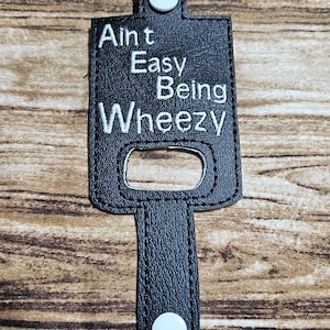 May include: Black leather keychain with a white embroidered saying "Ain't Easy Being Wheezy". The keychain has a metal loop and a white button on the bottom.