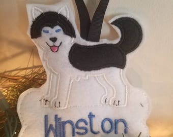 May include: White felt ornament with a black and white husky dog design. The dog has blue eyes and a pink tongue. The ornament has a bone shape with the name "Winston" embroidered in blue thread.