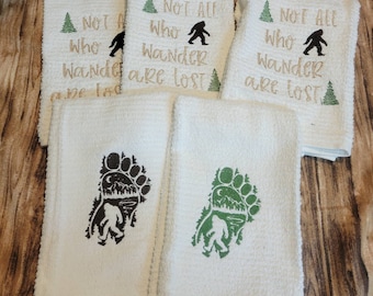 May include: White hand towels with embroidered designs. Some towels feature the text "NOT ALL WHO WANDER ARE LOST" with a Bigfoot silhouette and a green tree. Other towels have a Bigfoot footprint design in brown and green.