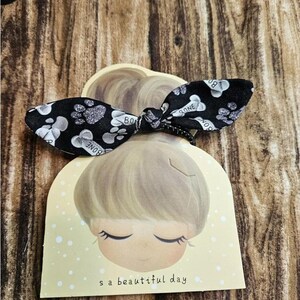 May include: Black and white hair tie with a paw print and bone design. The hair tie is tied in a bow and is sitting on a yellow card with a cartoon girl and the text "It's a beautiful day".