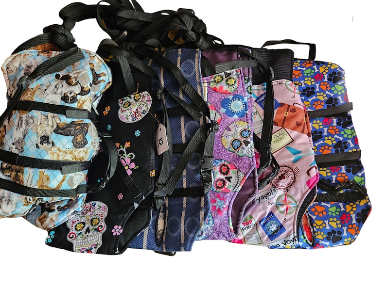May include: Six different dog harnesses with various colorful patterns, including skulls, paw prints, and travel-themed designs. The harnesses are made of fabric and have adjustable straps.