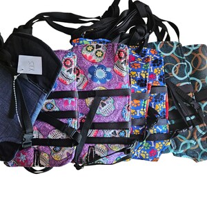 May include: Five different dog harnesses with adjustable straps. The harnesses are made of fabric and have various patterns, including skulls, paw prints, and circles. One harness is solid blue with the text "SXS" on it.