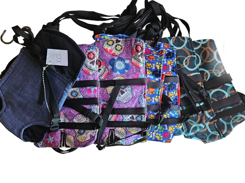 May include: Five different dog harnesses with adjustable straps. The harnesses are made of fabric and have various patterns, including skulls, paw prints, and circles. One harness is solid blue with the text "SXS" on it.