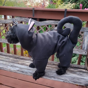 May include: A black dog wearing a black cat costume with pink ears and a curled tail. The dog is sitting on a wooden bench.