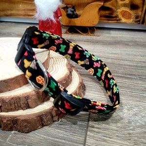 Puede incluir: Un collar de perro negro con un estampado navide&ntilde;o festivo que incluye galletas de jengibre, bastones de caramelo y copos de nieve. El collar tiene una hebilla negra y un anillo en D plateado.