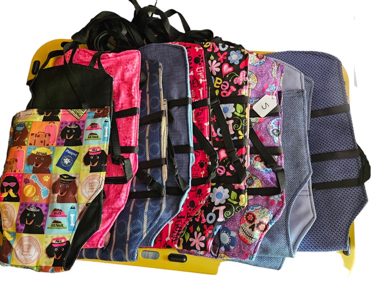 May include: A collection of colorful dog harnesses with various patterns, including a dachshund print, a floral print, and a skull print. The harnesses have adjustable straps and are made of durable fabric.