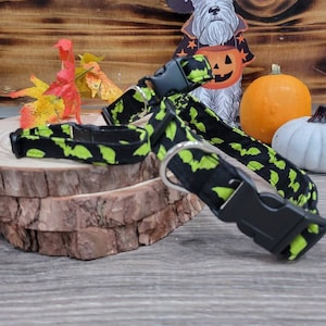 May include: A black and green dog collar with a bat pattern. The collar has a black plastic buckle and is on a wooden surface.