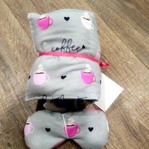 May include: A gray and pink coffee-themed eye mask and a matching blanket. The blanket is folded and tied with a pink ribbon. The eye mask is shaped like a butterfly. Both items have a pattern of pink coffee cups with steam and black hearts.