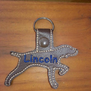May include: Brown leather dog-shaped keychain with a silver ring and the name "Lincoln" embroidered in blue thread.