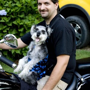 May include: A man wearing a black t-shirt and a black baseball cap is holding a small gray and white dog in a blue and black carrier. The man is sitting on a black motorcycle.