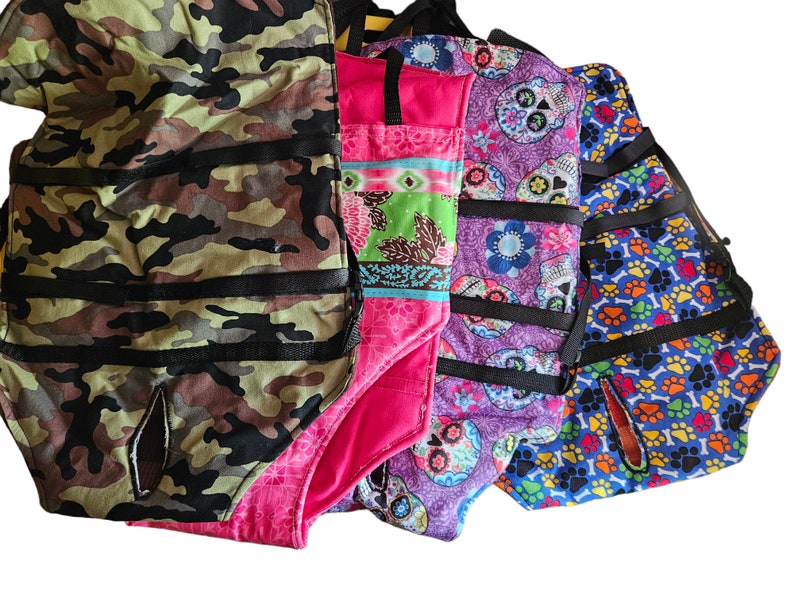 May include: Four different fabric dog harnesses with various patterns. The harnesses are made of a soft, durable fabric and feature adjustable straps for a comfortable fit. The patterns include camouflage, pink floral, purple skulls, and blue paw prints.
