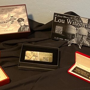 May include: Four wooden boxes with engraved portraits and QR codes. The boxes are on a black fabric surface. The text on the boxes reads "Robert Jacobson 180", "In Loving Memory Of Lou Witzeman", "Joseph Wojcik 77", and "Rural/Metro Ambulance Services".