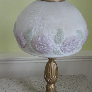 May include: A white table lamp with a floral design on the shade. The shade is made of glass and has a textured surface. The base of the lamp is gold metal and has a decorative design.