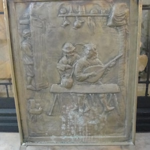 May include: A bronze-colored fireplace screen with a detailed relief depicting a medieval scene. The artwork shows figures playing music and other activities. The screen has a rectangular frame and two curved legs.