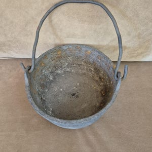 May include: A round, gray cast iron cauldron with a textured, weathered surface showing signs of oxidation and rust. The vessel features a thick, arched metal handle attached to two side loops, providing a sturdy grip for this heavy cooking pot.