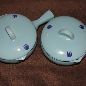 Könnte beinhalten: Zwei hellblaue Keramik-Ramekins mit blauen Blumendesigns. Jedes Ramekin hat einen Griff und einen Deckel.