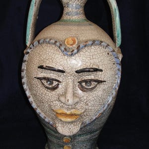 Peut inclure: Une grande cruche en céramique blanc cassé avec un visage. La cruche comporte deux anses et un visage en forme de cœur avec des yeux, un nez et une bouche. La cruche a un glaçage craquelé et est décorée d'accents bleus, verts et jaunes.