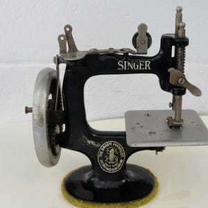 Puede incluir: Una máquina de coser manual Singer negra vintage con un volante de metal plateado y una placa base plana. La máquina presenta el nombre de la marca Singer en letras blancas y un logotipo circular que dice The Singer Manfg. Co. Trade Mark.