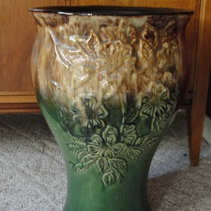 May include: A large, green and brown ceramic planter with a floral design. The planter has a wide base and a narrow neck.