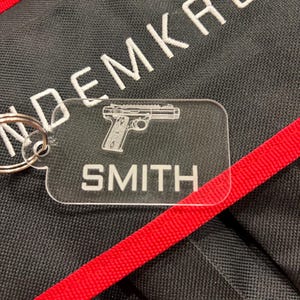 May include: Clear acrylic keychain with a silver keyring. The keychain features a detailed outline of a handgun and the name "SMITH" in white lettering. The background is black with white embroidered text and a red strap.