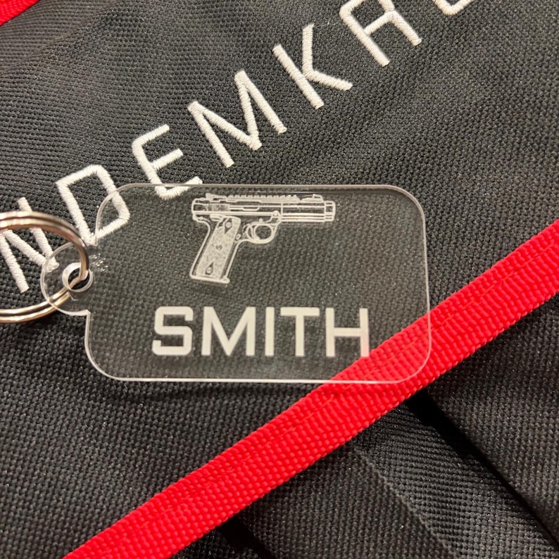 May include: Clear acrylic keychain with a silver keyring. The keychain features a detailed outline of a handgun and the name "SMITH" in white lettering. The background is black with white embroidered text and a red strap.