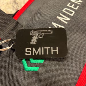 May include: Black rectangular keychain with a silver gun graphic and the name "SMITH" in white. The keychain is attached to a silver ring. The background is a black and gray fabric with white text and red trim.