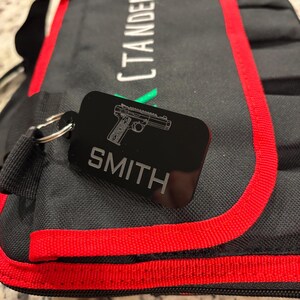 May include: Black rectangular keychain with a silver gun graphic and the name "SMITH" engraved in silver. The keychain is attached to a black bag with red trim. The bag has the word "CTANDER" printed on it.