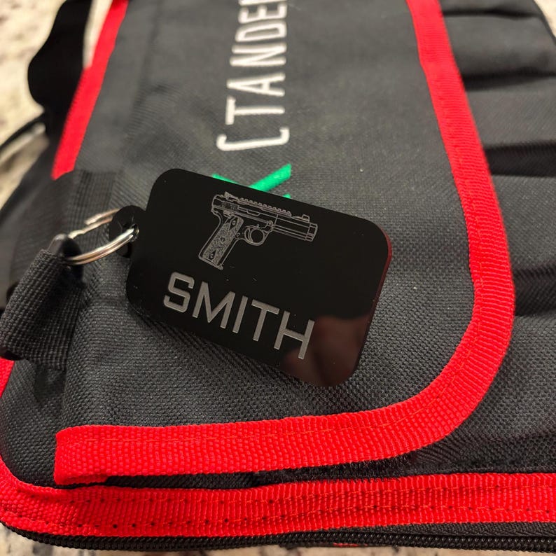 May include: Black rectangular keychain with a silver gun graphic and the name "SMITH" engraved in silver. The keychain is attached to a black bag with red trim. The bag has the word "CTANDER" printed on it.