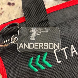 May include: A clear acrylic keychain with a silver keyring. The keychain features a detailed outline of a handgun and the name "ANDERSON" in a bold, sans-serif font. Below the name are three green chevrons. The background is a black bag with red trim.