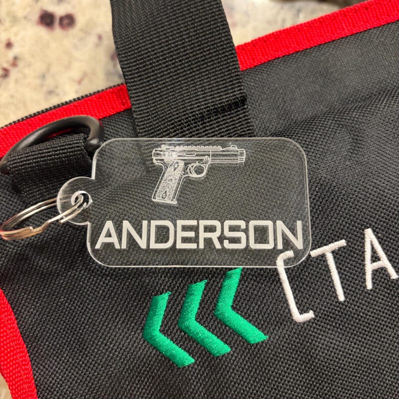 May include: A clear acrylic keychain with a silver keyring. The keychain features a detailed outline of a handgun and the name "ANDERSON" in a bold, sans-serif font. Below the name are three green chevrons. The background is a black bag with red trim.