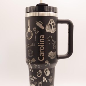 Pu&ograve; includere: Tazza da viaggio nera con manico e cannuccia, con icone a tema fitness incise e il nome "Carolina". La tazza include immagini di manubri, kettlebell e una borsa da palestra. &Egrave; visibile anche il testo "Sore Today, Strong Tomorrow".