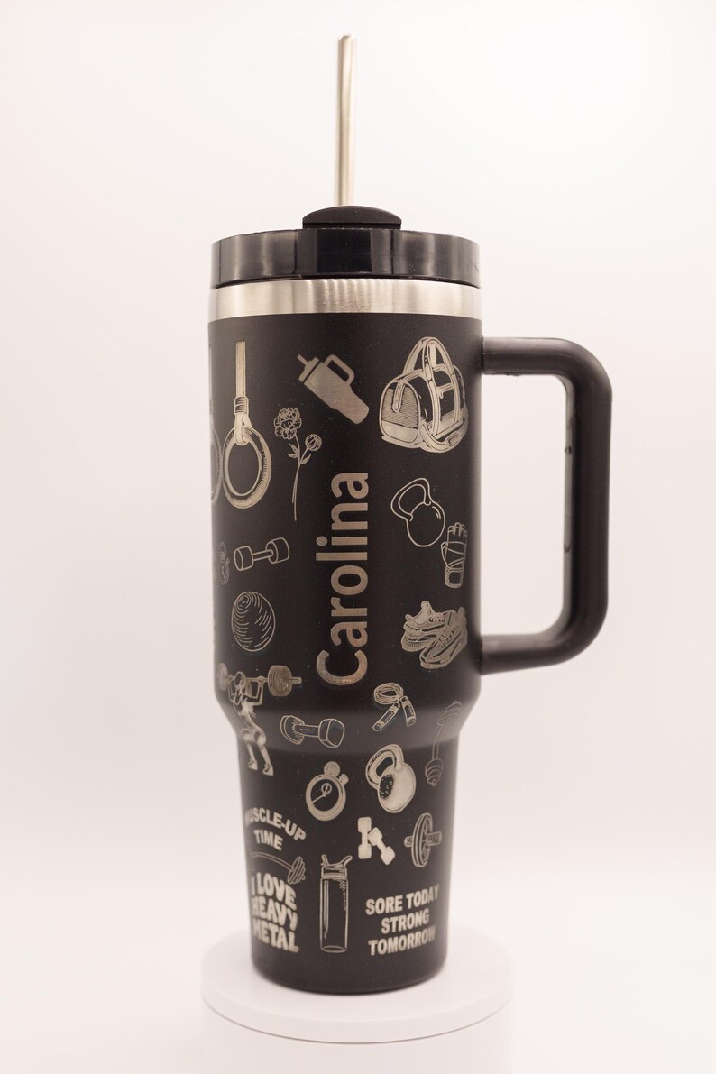 Pu&ograve; includere: Tazza da viaggio nera con manico e cannuccia, con icone a tema fitness incise e il nome "Carolina". La tazza include immagini di manubri, kettlebell e una borsa da palestra. &Egrave; visibile anche il testo "Sore Today, Strong Tomorrow".