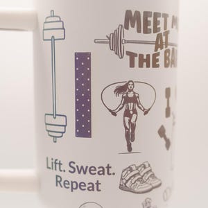 Pu&ograve; includere: Tazza da viaggio bianca con bordo arcobaleno e manico. La tazza presenta illustrazioni a tema fitness, tra cui un bilanciere, una donna che salta la corda e il testo "Meet Me At The Bar" e "Lift. Sweat. Repeat."