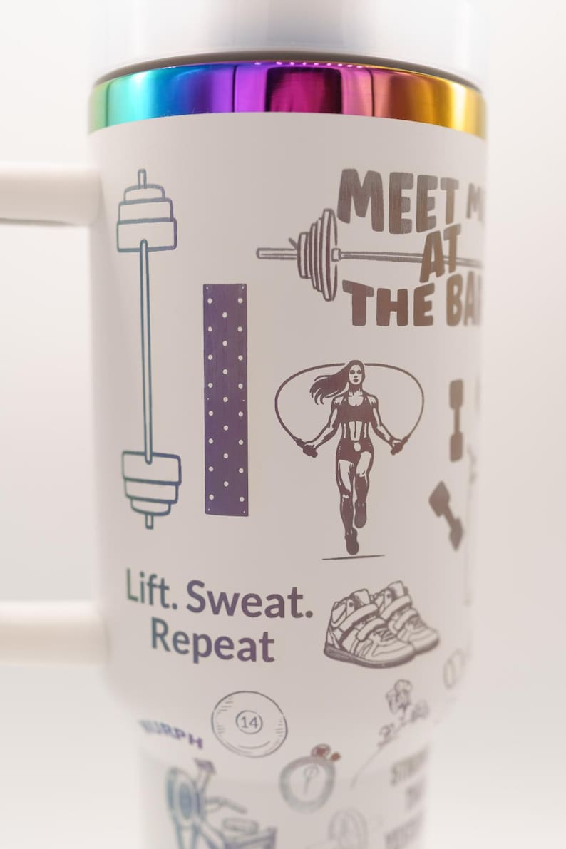 Pu&ograve; includere: Tazza da viaggio bianca con bordo arcobaleno e manico. La tazza presenta illustrazioni a tema fitness, tra cui un bilanciere, una donna che salta la corda e il testo "Meet Me At The Bar" e "Lift. Sweat. Repeat."