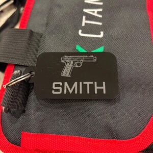 May include: Black rectangular keychain with a silver outline of a handgun and the name "SMITH" in capital letters. The keychain is attached to a silver keyring. The background is a black and red bag.
