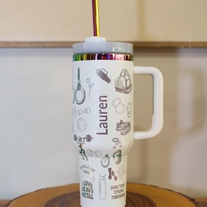 Pu&ograve; includere: Bicchiere bianco con manico, coperchio e cannuccia arcobaleno. Il bicchiere &egrave; decorato con illustrazioni a tema fitness e il nome "Lauren". Il bicchiere &egrave; appoggiato su una superficie di legno.