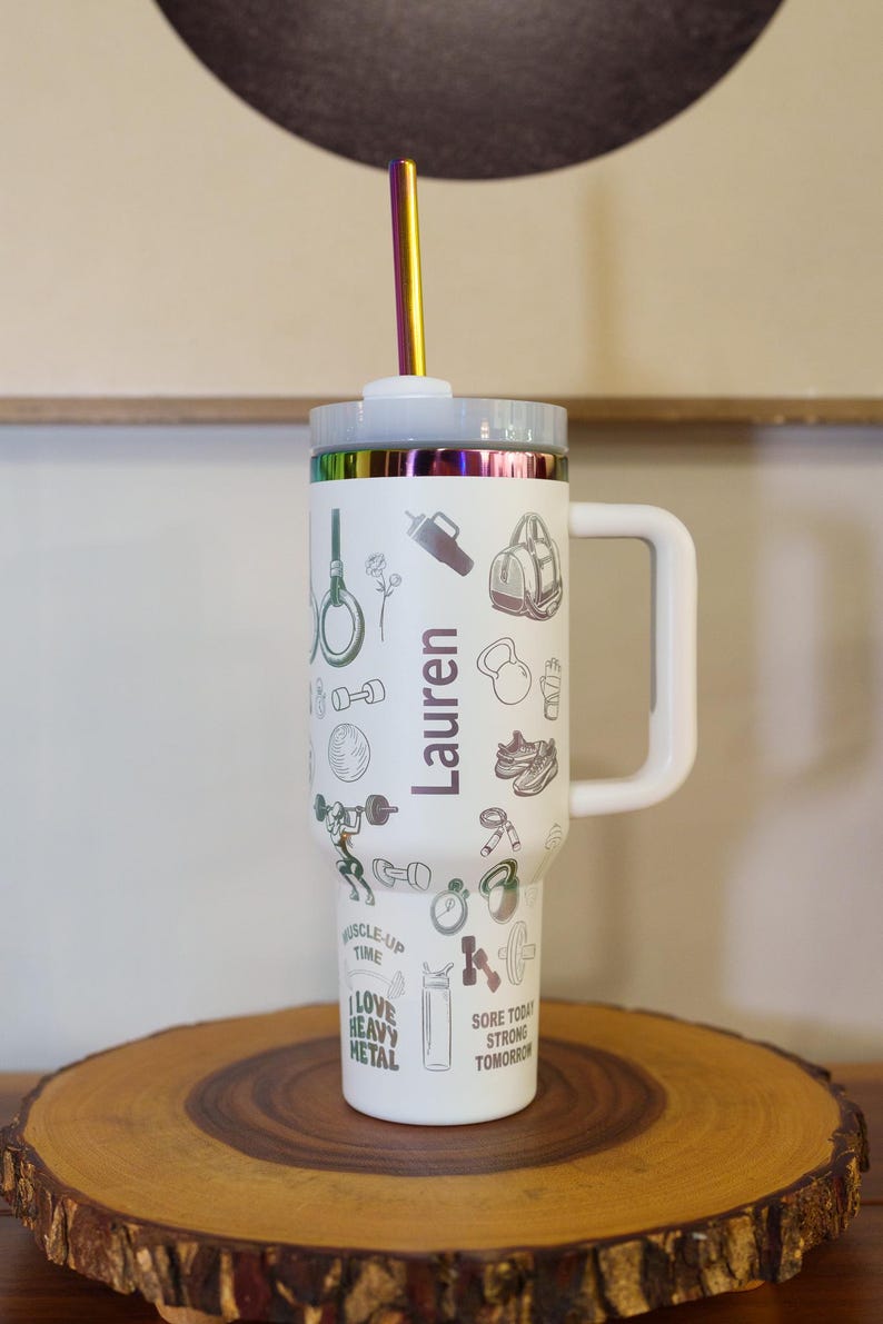 Pu&ograve; includere: Bicchiere bianco con manico, coperchio e cannuccia arcobaleno. Il bicchiere &egrave; decorato con illustrazioni a tema fitness e il nome "Lauren". Il bicchiere &egrave; appoggiato su una superficie di legno.