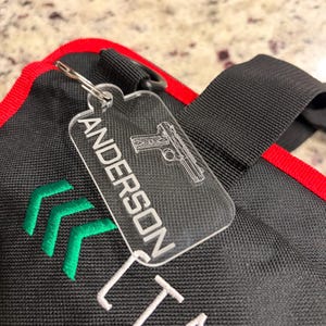 May include: Clear acrylic keychain with the name "ANDERSON" and a gun design. The keychain is attached to a black bag with red trim and green embroidered arrows.