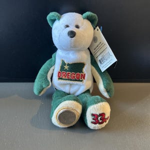 May include: A plush bear toy with a white head and torso, green arms, ears, and legs. The bear has a black nose and the state of Oregon is printed on the torso. The bear's foot has the number 33 printed on it.
