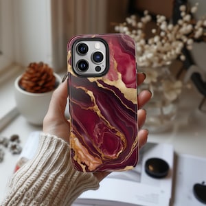 May include: A phone case with a marble design in shades of burgundy, gold, and white. The case has a raised camera bump and a cutout for the phone's charging port.