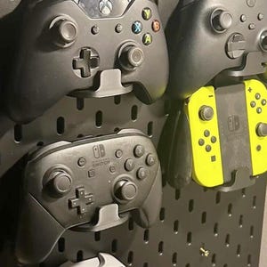 May include: A black pegboard with several video game controllers mounted on it. Controllers include Xbox and Nintendo Switch models. The Switch controllers are yellow. The pegboard is designed for game console organization.