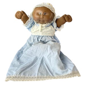 Pode incluir: Uma boneca Cabbage Patch Kid vintage com pele marrom, olhos castanhos e um vestido azul com renda branca. A boneca está usando uma touca branca com renda.