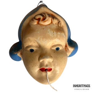 Puede incluir: Máscara vintage envejecida de una cara de niño, con pelo y ojos azules y labios rojos. Una cuerda sale de la boca. La máscara tiene un aspecto envejecido y antiguo, con desgaste visible. La máscara probablemente está hecha de un material similar al yeso.