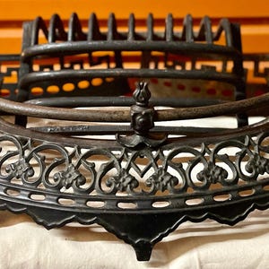 May include: Black cast iron fireplace grate with an ornate design. The grate has a curved top and a decorative bottom edge.