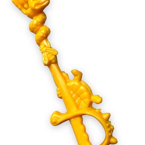 Puede incluir: Una espada de juguete de plástico amarillo con una serpiente, una tortuga y otros elementos decorativos. La espada tiene un mango con una guarda con púas.