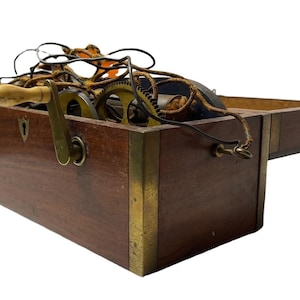 Puede incluir: Una caja de madera vintage con esquinas de latón y tapa con bisagras. En el interior hay engranajes, cables y una manivela. La caja tiene un ojo de cerradura y un asa de madera. La combinación de colores general es marrón y dorado.