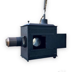 Puede incluir: Un proyector de diapositivas vintage de metal negro con una lente y una puerta que se abre para revelar el interior. El proyector tiene un asa en la parte superior y una pequeña perilla de metal en el lateral.