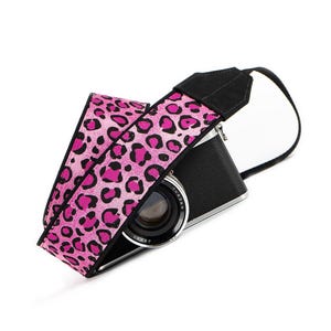 May include: A black camera with a lens attached to a wide camera strap featuring a vibrant pink and black leopard print pattern. The strap includes black suede-like end tabs that connect to the camera body, creating a functional accessory.