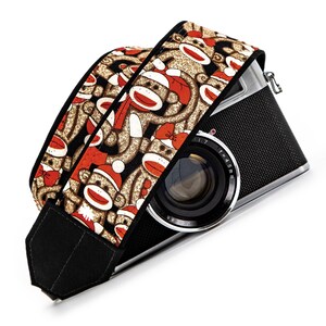 May include: A black camera with a patterned camera strap. The strap features a repeating design of sock monkeys in red, white, and brown on a black background. The camera lens is visible.