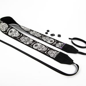 Black and white camera strap with a repeating pattern of sugar skulls. The skulls are intricately detailed with floral designs. The strap is made of a durable fabric and features a comfortable, adjustable design.  Perfect for photographers who appreciate unique and stylish accessories.